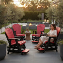 A woman reads on a Kanyon Living Poly Lumber Adirondack Glider Chair Tall in red and black, surrounded by matching chairs. String lights hang above and a fire pit glows at sunset, showcasing the eco-friendly furniture.