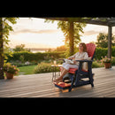 A woman relaxes on a red Adirondack chair on a wooden patio at sunset, holding a book. The patio overlooks a lush garden with blooming flowers and greenery, with a drink nearby on a small table.