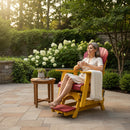 A woman sits on a Kanyon Living Poly Lumber Adirondack Glider Chair Tall Red and Yellow, holding a cup with a white blanket, beside a small table with a potted plant in a flowering garden with stone walls on a sunny day.