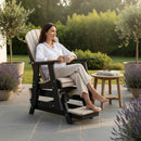 A woman relaxes barefoot in the Kanyon Living Poly Lumber Adirondack Glider Chair Tall, Seashell and Black, on a stone patio with potted plants, a small table, and books beside her, enjoying a peaceful sunny day.