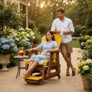 A woman in a light blue dress relaxes on a Kanyon Living Poly Lumber Adirondack Glider Chair Tall Yellow and Cedar Wood on the patio, surrounded by hydrangeas, while a man in a white shirt stands behind her with coffee. A table with a drink is nearby.