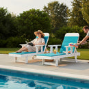 A woman relaxes on the Kanyon Living Poly Lumber Chaise Lounge with Arms in Aruba Blue & White by the pool, reading a book, as a man sets down towels. Drinks and snacks are on a table between them, surrounded by lush greenery.