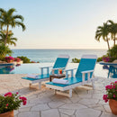 Two Kanyon Living Poly Lumber Chaise Lounges with arms in Aruba Blue & White, topped with rolled towels, face an infinity pool and ocean at sunset on a stone patio surrounded by palm trees and pink flowers, with a small table and drinks between them.