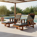 Two Kanyon Living Poly Lumber Chaise Lounges with Arms in Black and Deluxe Antique Mahogany sit poolside under a pergola, rolled white towels on top. A small table holds drinks, surrounded by lush trees and shrubs for a relaxing outdoor retreat.