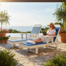 A woman in a sun hat and white dress relaxes by the pool on a Kanyon Living Poly Lumber Chaise Lounge with Arms in Blue and Light Grey, reading a book. A drink, towel, and flowers rest nearby amid ocean views and lush greenery.