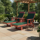 Two Kanyon Living Poly Lumber Chaise Lounges with Arms in Dark Green and Cherry Wood sit side by side beneath a pergola, surrounded by lush greenery and potted plants. A small round table holds two glasses of lemonade.