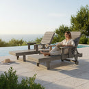 A woman in light clothing relaxes by the pool on a Kanyon Living Poly Lumber Chaise Lounge with Arms Deluxe Driftwood Grey, reading a book. An empty chair and sun hat rest nearby, while the sea glimmers beyond this eco-friendly furniture.