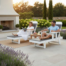 A woman and man relax outdoors on Kanyon Living Poly Lumber Chaise Lounges with Arms Deluxe in Driftwood Grey and White, near a table with fruit and drinks, surrounded by greenery, flowers, a fireplace, and trees at sunset.