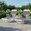 Two Kanyon Living Poly Lumber Chaise Lounges with Arms Deluxe in Driftwood Grey and White rest on a stone patio, with a clear tray holding two glasses between them, surrounded by blooming hydrangeas—ideal for outdoor relaxation.