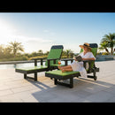 A woman in a sunhat and white dress relaxes by the pool on a Kanyon Living Poly Lumber Chaise Lounge with Arms in Lime Green and Black, reading a magazine, with a drink nearby under palm trees and clear, sunny skies.