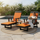 A woman in a white dress relaxes by the pool, reading on a Kanyon Living Poly Lumber Chaise Lounge with Arms in Orange and Black. Nearby, drinks and a hat rest on eco-friendly furniture atop a round rug with greenery behind.