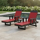 Two Kanyon Living Poly Lumber Chaise Lounges with Arms in Red and Black are placed poolside, separated by a small table with refreshments. Surrounded by greenery and flowers, this Amish-crafted outdoor furniture adds style to any setting.