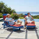 A woman in a sunhat and a man in sunglasses relax on red and blue loungers by a pool. A small table with drinks and fruit is between them. Trees and water are in the background under a clear sky.
