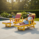 A woman in a light blue dress and sunhat relaxes poolside, reading on a Kanyon Living Poly Lumber Chaise Lounge with Arms in red and yellow. An empty chair and side table with a drink sit nearby, surrounded by greenery and flowers.