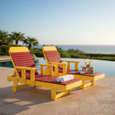 Two Kanyon Living Poly Lumber Chaise Lounges with Arms in red and yellow sit poolside, facing the ocean. A matching side table holds snacks and drinks, while rolled towels rest on a chair. The area is surrounded by palm trees and greenery.