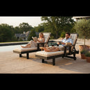 A woman reads and a man eats as they relax, smiling, on Kanyon Living Poly Lumber Chaise Lounges with Arms in Seashell and Black by the pool, surrounded by potted plants and trees in a sunny outdoor setting.