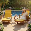 A woman in a white dress relaxes by the pool on a Kanyon Living Poly Lumber Chaise Lounge with Arms in Yellow and Cedar Wood, sipping a drink. Nearby, a table holds a pitcher and glass, surrounded by lush greenery and blooming flowers.