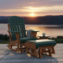 A green Adirondack rocking chair with a blanket rests on a lakeside deck at sunset beside an Amish-crafted Kanyon Living Poly Lumber Gliding Ottoman in Dark Green and Cedar Wood, paired with a small table holding a steaming mug.