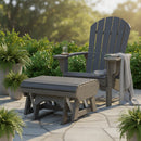 A Kanyon Living Poly Lumber Gliding Ottoman Deluxe in Driftwood Grey pairs with an Adirondack chair on a stone patio with potted ferns, a striped blanket, iced tea, and lush greenery completing the outdoor scene.