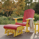 A red and yellow Kanyon Living Poly Lumber Adirondack chair with a matching Kanyon Living Gliding Ottoman sits on a wooden deck. A beige blanket hangs over the armrest, and a mug rests on a small yellow table amid lush green plants.