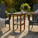 A cozy patio features two dark Adirondack chairs and the Kanyon Living Poly Lumber Tall Oval Side Table in Black and Deluxe Antique Mahogany, holding eyeglasses and a mug. A person sits on the right chair, with string lights and greenery in the background.