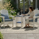 A woman sits on a patio chair with blue cushions, reaching for a steaming mug on the Kanyon Living Poly Lumber Tall Oval Side Table in blue and light grey. Another chair and potted plant rest on a patterned outdoor rug in the sunny garden.