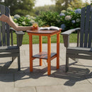 A Kanyon Living Poly Lumber Tall Oval Side Table in Dark Wood and Orange sits between two gray chairs on a sunny patio, holding a drink, book, and glasses as someone reaches for their beverage surrounded by garden flowers.