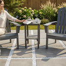 A woman sits on a gray chair beside a Kanyon Living Poly Lumber Tall Oval Side Table Deluxe in Driftwood Grey with a magazine, mug, and teapot; another empty chair is nearby on a stone patio surrounded by green grass and garden plants.