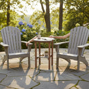 Two empty Adirondack chairs face each other on a stone patio, with the Kanyon Living Poly Lumber Tall Oval Side Table in Light Grey and Weathered Wood between them. Coffee mugs and a book rest atop it, framed by lush greenery and flowers.