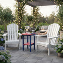 Two white Adirondack chairs and a small blue-and-red table sit on a stone patio surrounded by blooming flowers, with a fire pit and plush seating in the background under a pergola. A pitcher and glass of lemonade are on the table.