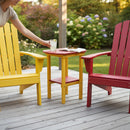 A person sets a glass of lemonade on the Kanyon Living Poly Lumber Tall Oval Side Table in red and yellow, surrounded by matching Adirondack chairs on a wooden deck. Sunglasses and an open book rest atop the table, with a lush garden as backdrop.