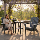 A woman reads in a blue Adirondack chair on a stone patio next to a Kanyon Living Poly Lumber Tall Oval Side Table in Seashell and Black, with another empty chair, fire pit, and blooming wisteria pergola nearby on a sunny day.