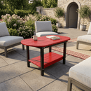 The Kanyon Living Poly Lumber Oval Coffee Table in red and black sits on a stone patio, surrounded by cushioned outdoor chairs and greenery, in front of a stone building with an arched doorway.