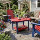 A vibrant patio scene features Kanyon Living’s Poly Lumber Oval Coffee Table in Red & Blue, placed among Adirondack chairs, drinks, plants, blooming flowers, stone planters, and a gray house as the backdrop.