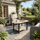 A cozy patio with eco-friendly wicker chairs and the Kanyon Living Poly Lumber Oval Coffee Table in Seashell & Black, holding a mug, plant, and book—surrounded by potted plants and flowers on a stone floor beneath a vine-covered pergola.