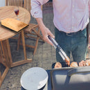 A person grills chicken outdoors with tongs. On the round wooden table are a plate, a glass of red wine, a brown paper bag, and the VIGOR Large Insulated Meat Resting Bag in the background.