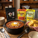 A hearty pot of chili made with the Meat Church Chili & Cornbread Comfort Bundle sits on a wooden table, surrounded by bowls, spoons, bags of original and jalapeño cornbread mix from Meat Church, and a basket of fresh baked cornbread squares.