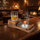 A glass of whiskey with ice and a cigar rest on the DDR BBQ Supply Multifunction Cigar & Whiskey Gift Set Stand with Ashtray and Drawer, atop a wooden tray in a cozy lounge with leather chairs, fireplace, and liquor shelves in the background.
