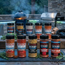 Ten bottles of Myron Mixon Ultimate BBQ Rub are displayed outdoors on a wooden surface with herbs, wood chunks, grills, and rising smoke in the background, capturing an authentic barbecue vibe and showcasing championship flavor.