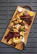 A Napoleon Charcuterie Board (70112) by Napoleon Grills with assorted grilled meats, veggies, and three ceramic ramekins rests on a gray table. A knife lies on the board featuring ribs, chicken, and skewered vegetables.