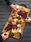 The Napoleon Grills Charcuterie Board (70112) displays assorted grilled meats, veggies, bread slices, and three sauce ramekins as two people reach for food on this acacia serving board set on a gray slatted table with utensils.