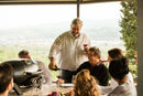 A smiling man pours red wine for an older man at an outdoor table, as friends enjoy a meal and cook together on the Napoleon TravelQ PRO285 Portable Gas Grill by Napoleon Grills with scenic views in the background.