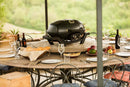A round outdoor table set for a meal with plates, glasses, wine bottles, fruit, and bread features the Napoleon TravelQ PRO285 Portable Gas Grill by Napoleon Grills at its center, surrounded by chairs and lush greenery.