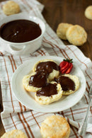 A plate with split biscuits topped with chocolate gravy sits on a striped cloth. A whole strawberry and extra biscuits are nearby, plus a bowl of Old School Brand Chocolate Gravy Mix—a classic Southern breakfast comfort from Old School Brand.