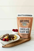A pouch of Old School Brand Chocolate Gravy Mix sits next to a plate of biscuits topped with chocolate gravy and fresh raspberries, offering a classic Southern breakfast. A small bowl of raspberries appears in the background.