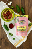 A bowl of Southern grits with sausage and jalapeños sits beside fresh jalapeños, a spoon, hot sauce, and a pink bag of Old School Brand Non-GMO Stone Ground White Unbleached Grits, 1lb on a wooden table.