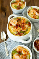 Four bowls of shrimp and Southern grits made with Old School Brand Non-GMO Stone Ground White Unbleached Grits are topped with bacon, herbs, green bell peppers, and sautéed shrimp, arranged on a gray-striped cloth with a spoon beside them.