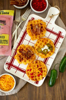 A white platter holds four savory waffles with bacon, sour cream with jalapeños, shredded cheese, and pimentos. Nearby are forks, jalapeños, grated cheese, pimentos, and a bowl of Old School Brand Non-GMO Stone Ground White Unbleached Grits.