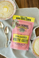 A bag of Old School Brand Non-GMO Stone Ground White Unbleached Grits, 1lb, sits on a gray cloth beside two bowls of white corn grits with butter and silver spoons on a wooden table.