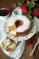 A moist cake made from Old School Brand Pound Cake Mix sits on a floral plate, dusted with powdered sugar. Two wedges rest nearby with berries, a knife, and a vase of red and purple flowers—classic old-fashioned flavor in every bite.